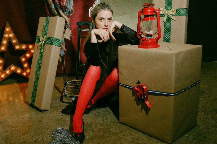 a girl with a white hair bow and red tights sits central to the image. She is surrounded by presents wrapped in brown paper.